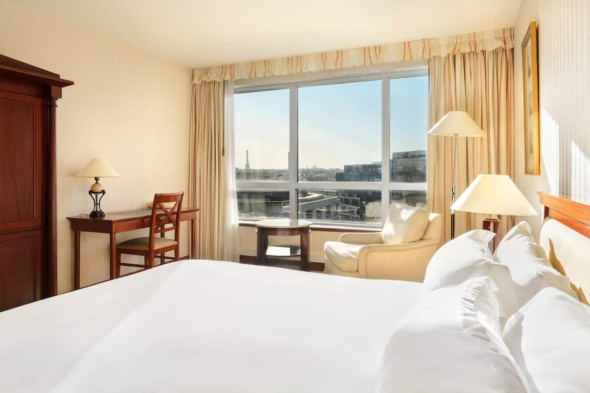 A hotel room with a large bed, a desk with a chair, a small table, and a view of the city through a large window.