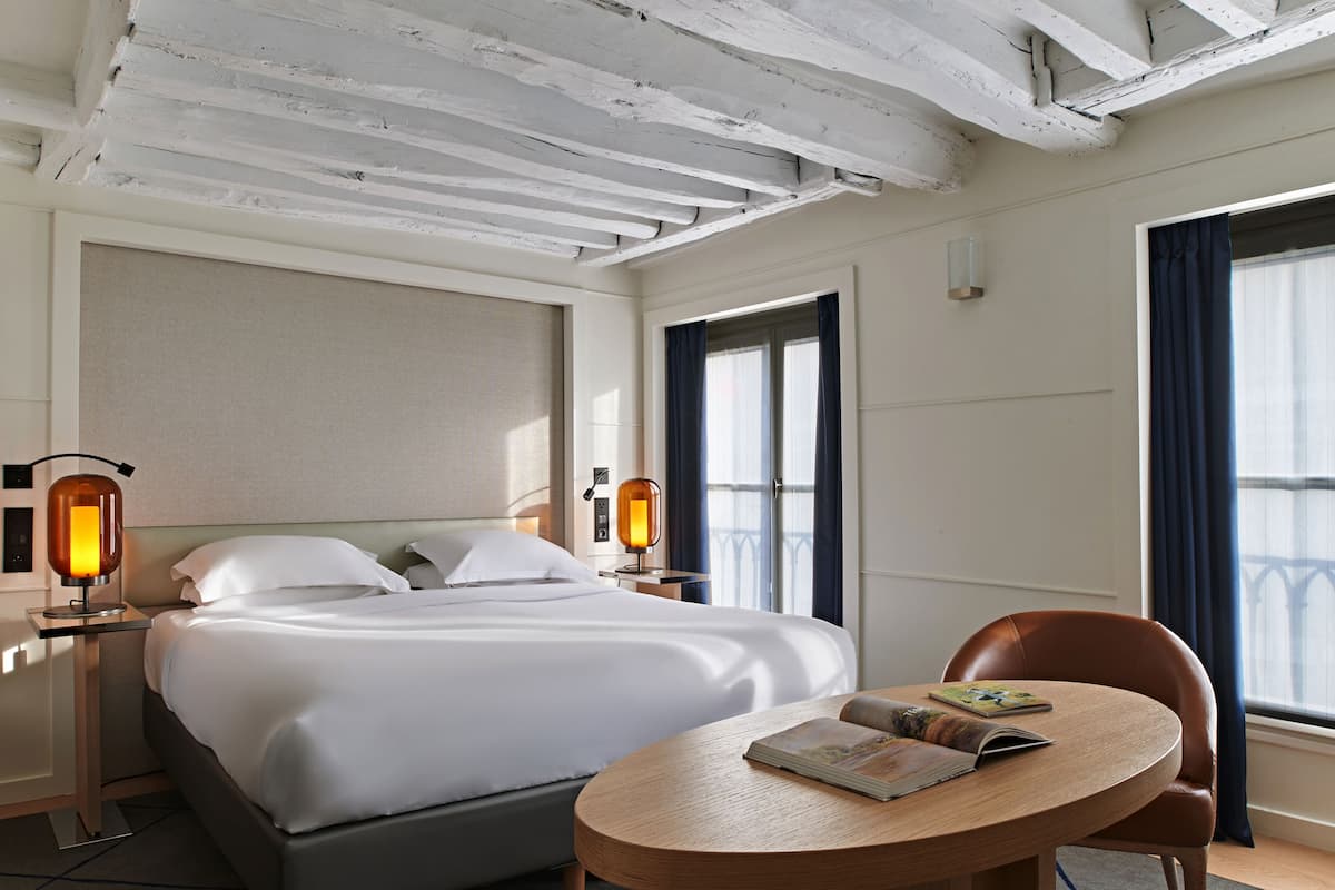 A hotel room with a bed, two bedside lamps, a round wooden table with a book, and a window with curtains.