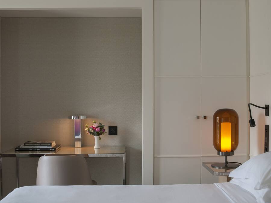 A hotel room with a bed, a desk with a lamp, a chair, and a small table with a vase of flowers.