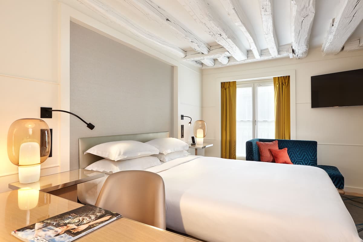 A hotel room with a large bed, a desk with a lamp, a chair, a TV mounted on the wall, and a window with curtains.