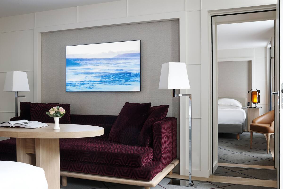 A modern hotel room with a large window, a sofa, a table with a book and a vase, and a bed with a nightstand and lamp.