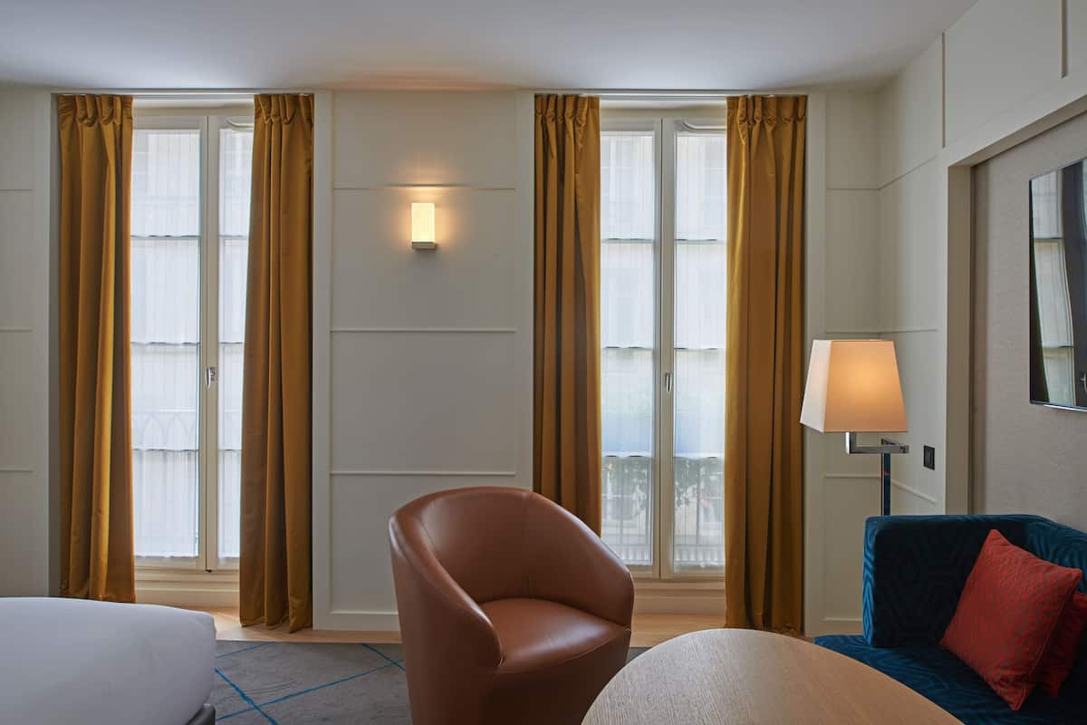 A hotel room with a bed, a chair, a sofa, a lamp, and large windows with curtains.