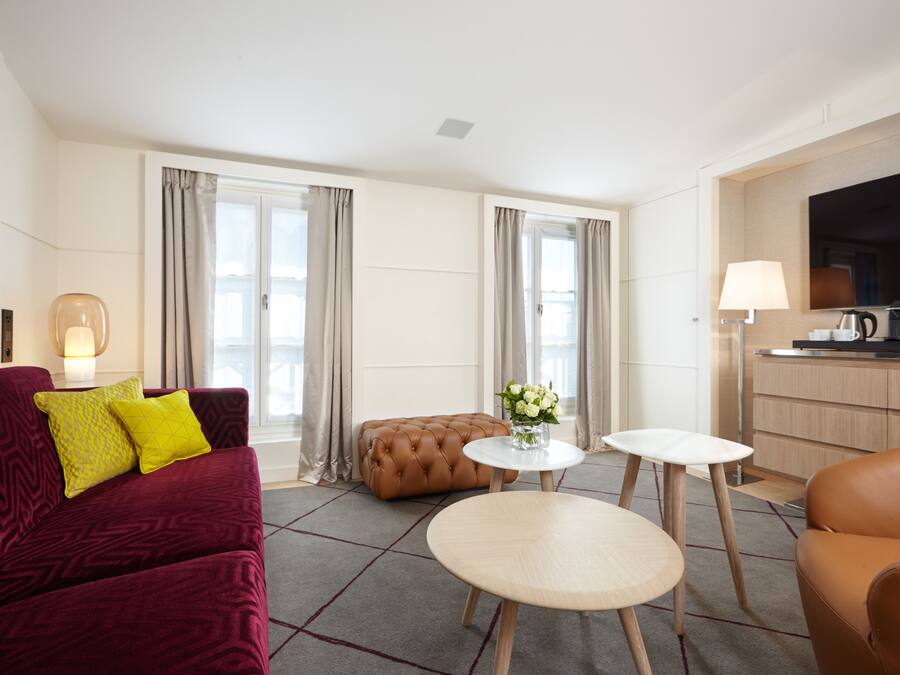 A modern hotel room with a red sofa, a brown ottoman, two round tables, a flat-screen TV, and a kitchenette.
