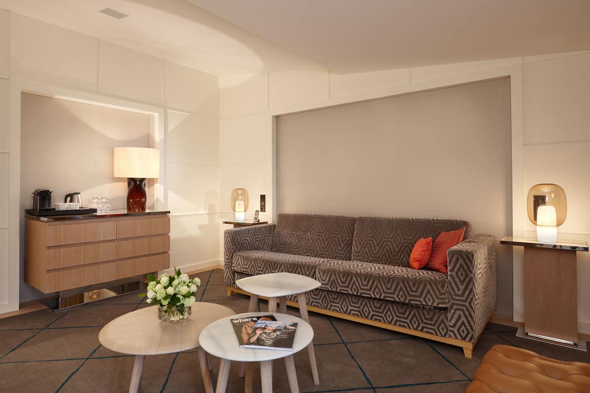 A modern hotel room with a sofa, two small tables, a coffee station, and a lamp.