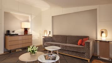 A modern hotel room with a sofa, two small tables, a coffee station, and a lamp.