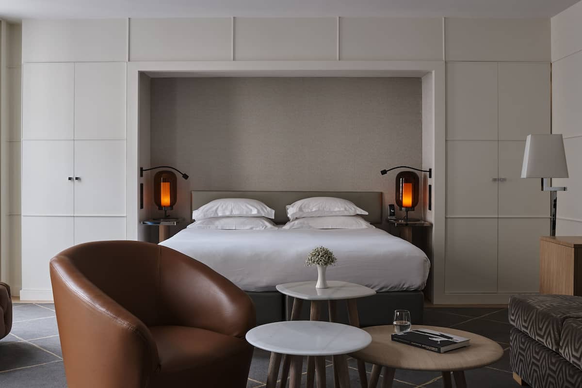 A hotel room with a large bed, two bedside tables, a small round table, a brown leather armchair, and a sofa.