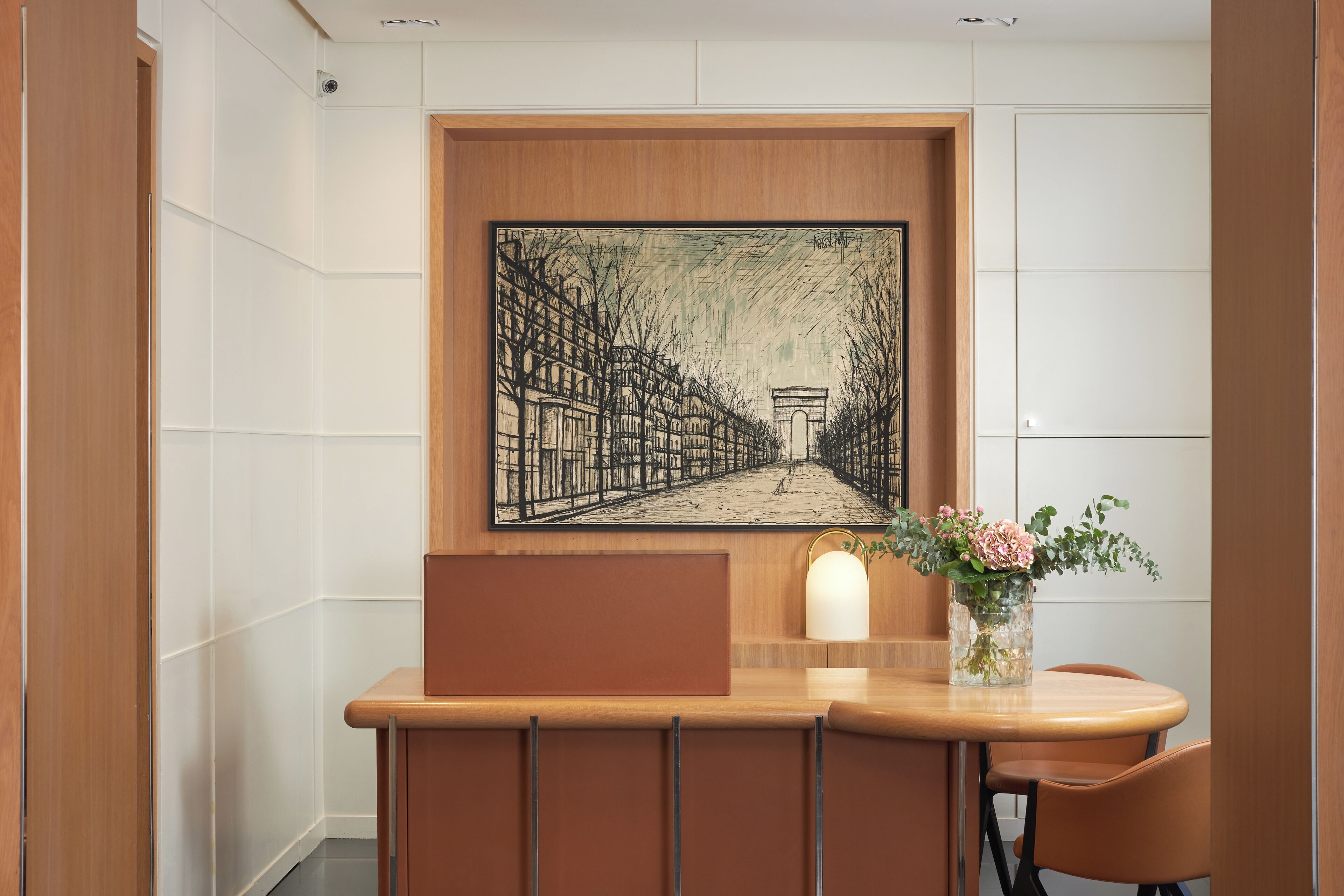A reception desk with a framed black and white street scene painting, a vase with flowers, and a wooden bench.