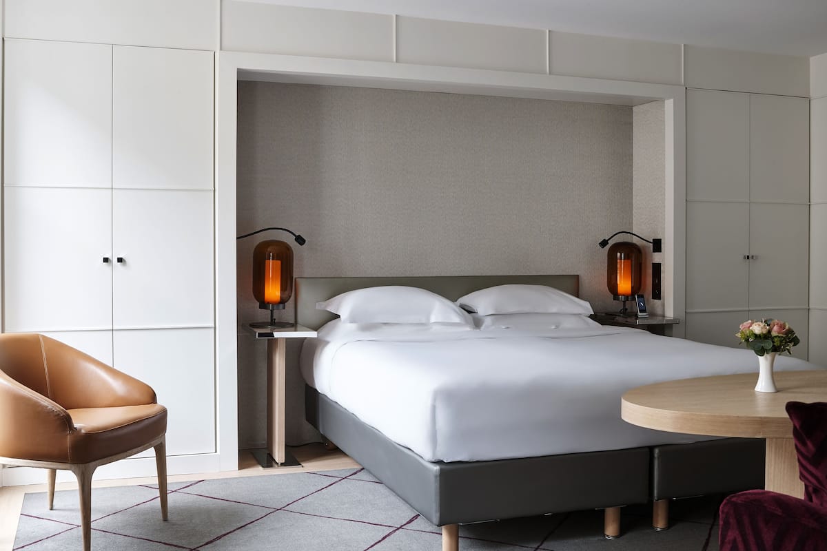 A modern hotel room with a large bed, two bedside tables with lamps, a chair, and a small round table with a vase of flowers.