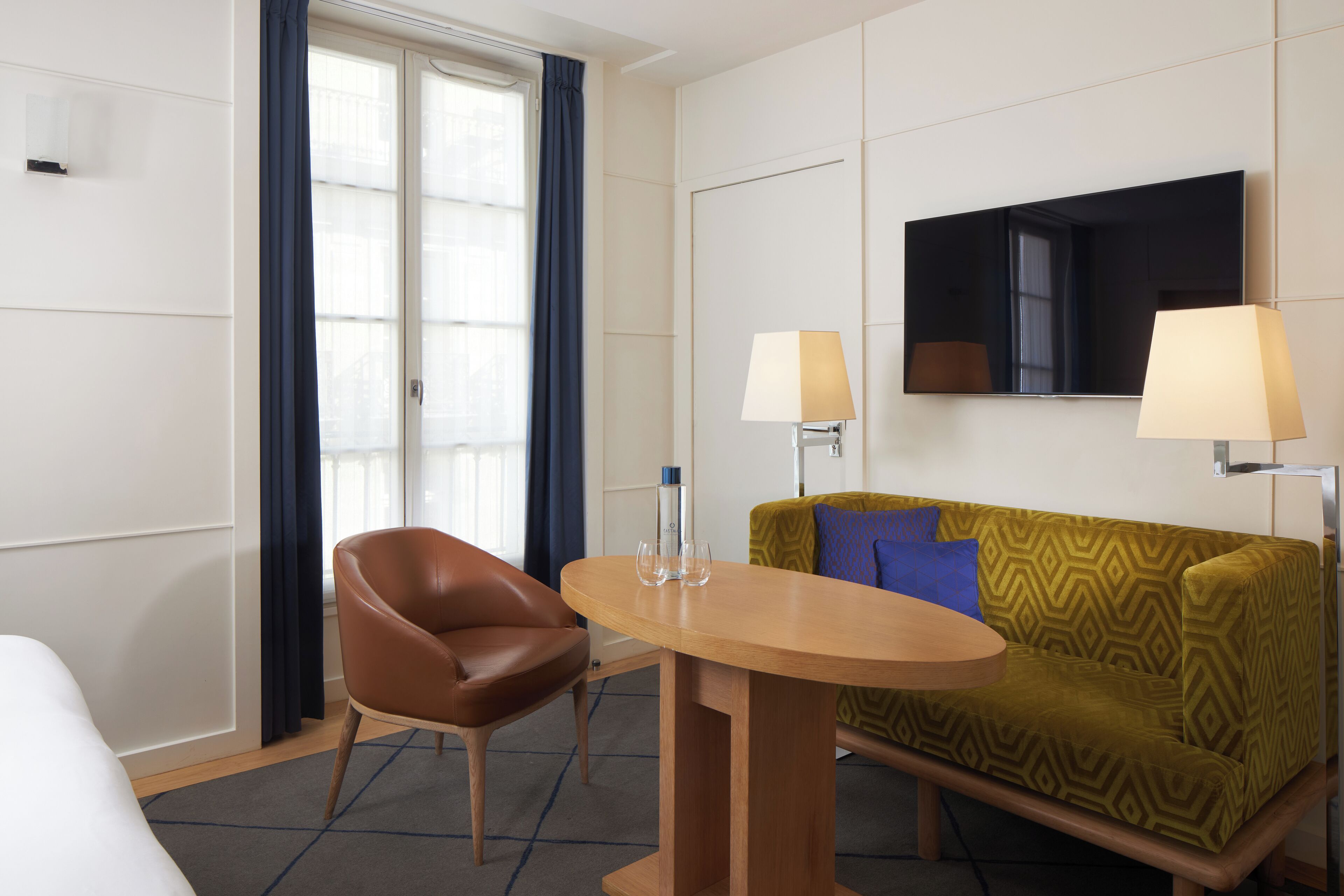 Une chambre d’hôtel avec un lit, un fauteuil en cuir marron, un canapé à motifs verts, une table en bois, deux lampes et un grand miroir au mur.