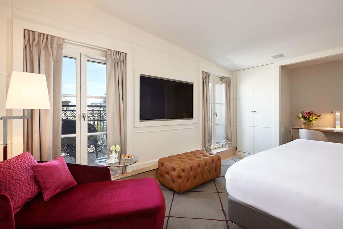 A hotel room with a large bed, a red sofa, a small table with a lamp, and a window with curtains.