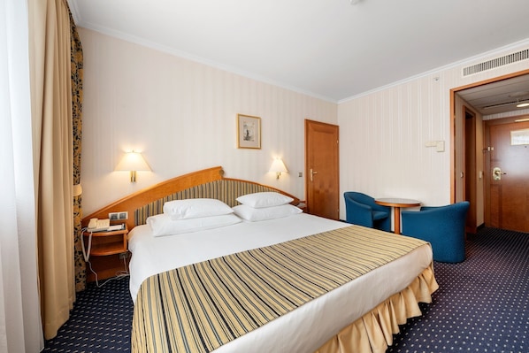 Habitación de hotel con una cama grande, lámparas de noche, una mesita, una silla y una puerta.
