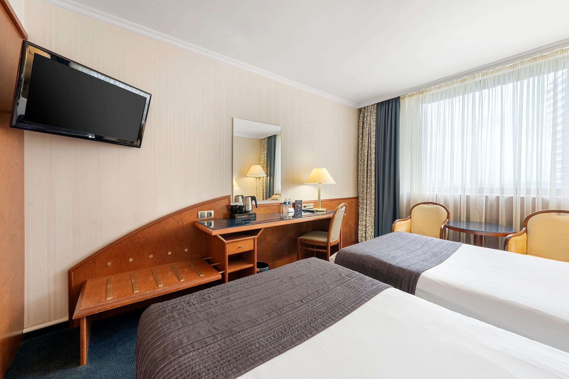 A hotel room with two beds, a desk, a chair, a TV, and a window with curtains.