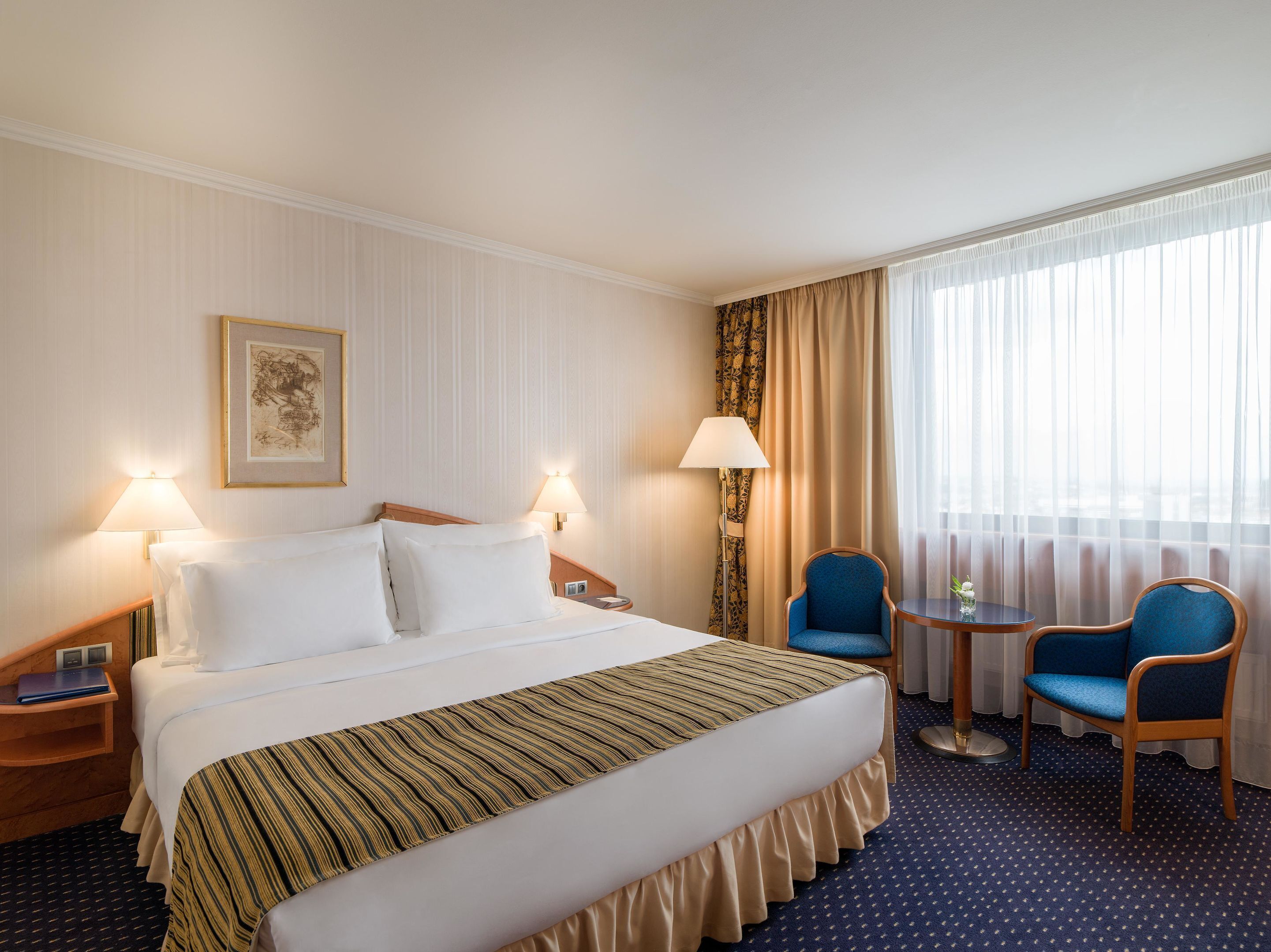 A hotel room with a large bed, two bedside lamps, a small table with a chair, and a window with sheer curtains.