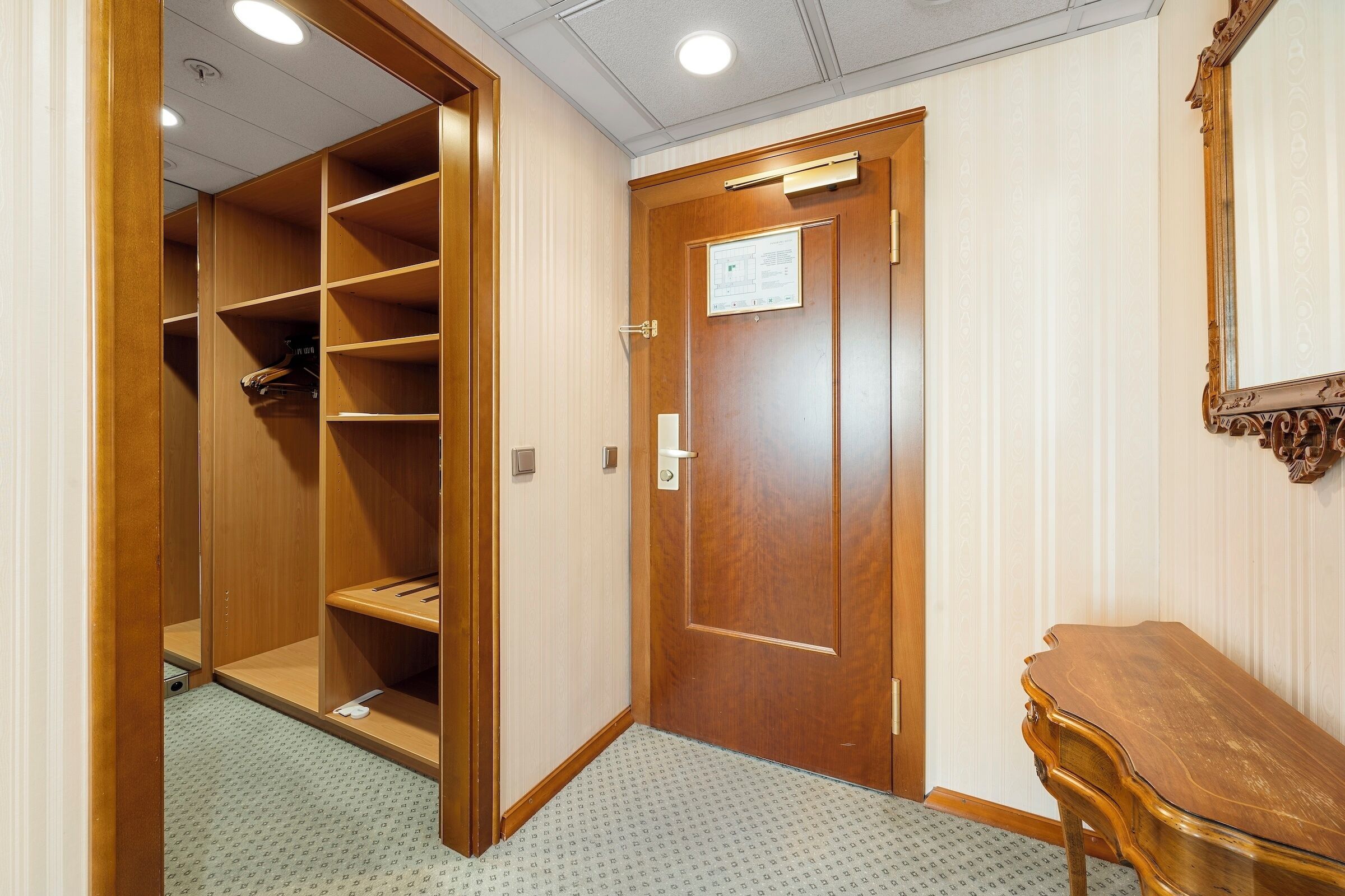 A hotel room with a wooden wardrobe, a small wooden table, and a door with a sign.