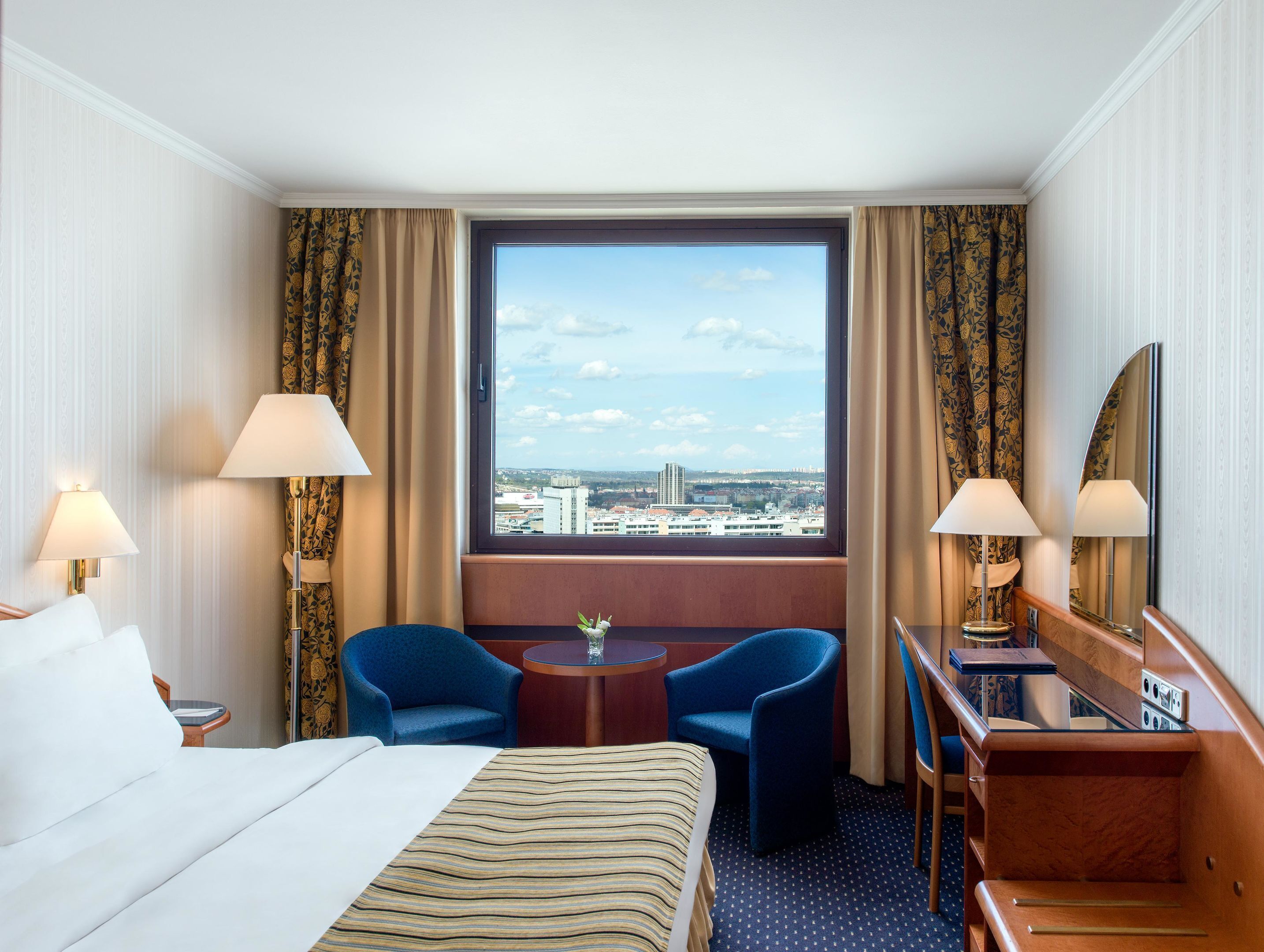 A hotel room with a bed, two chairs, a small table, a mirror, and a window with a city view.