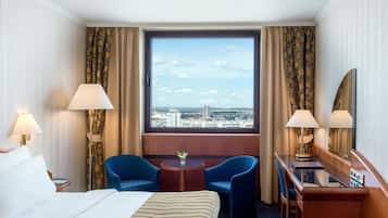 A hotel room with a bed, two chairs, a small table, a mirror, and a window with a city view.