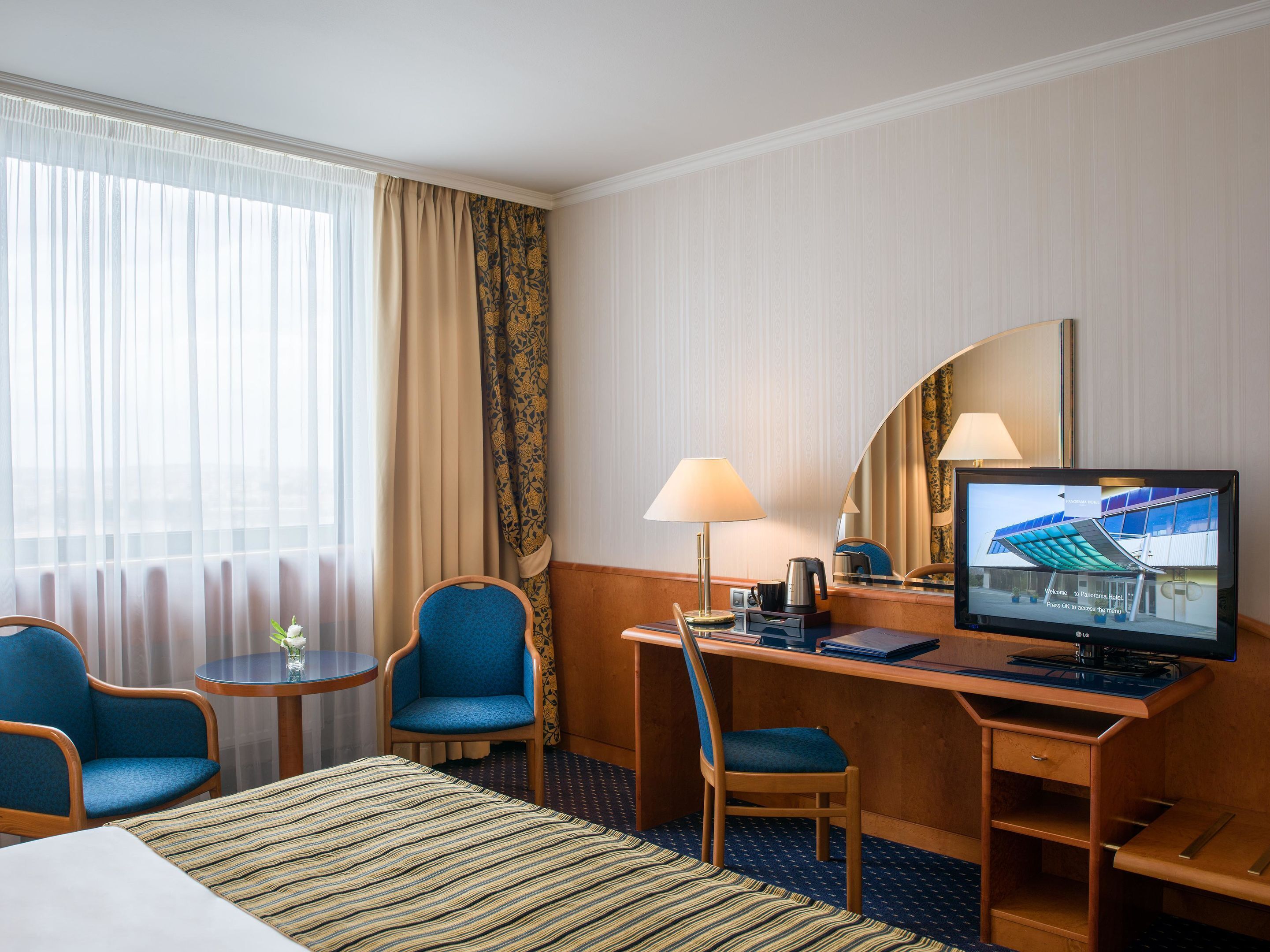 A hotel room with a bed, a desk, two chairs, a television, and a window with curtains.