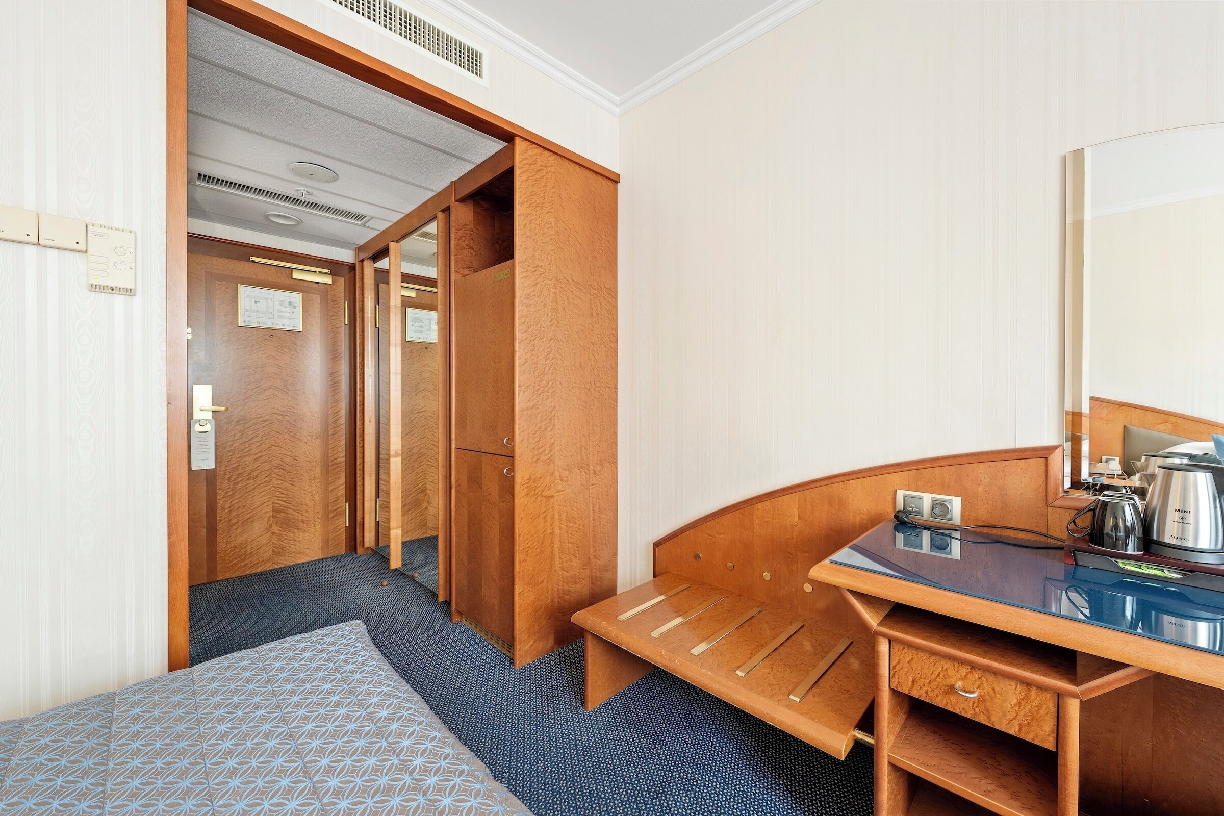 A compact hotel room with a bed, a wooden desk, a bench, a small drawer, and a large mirror.