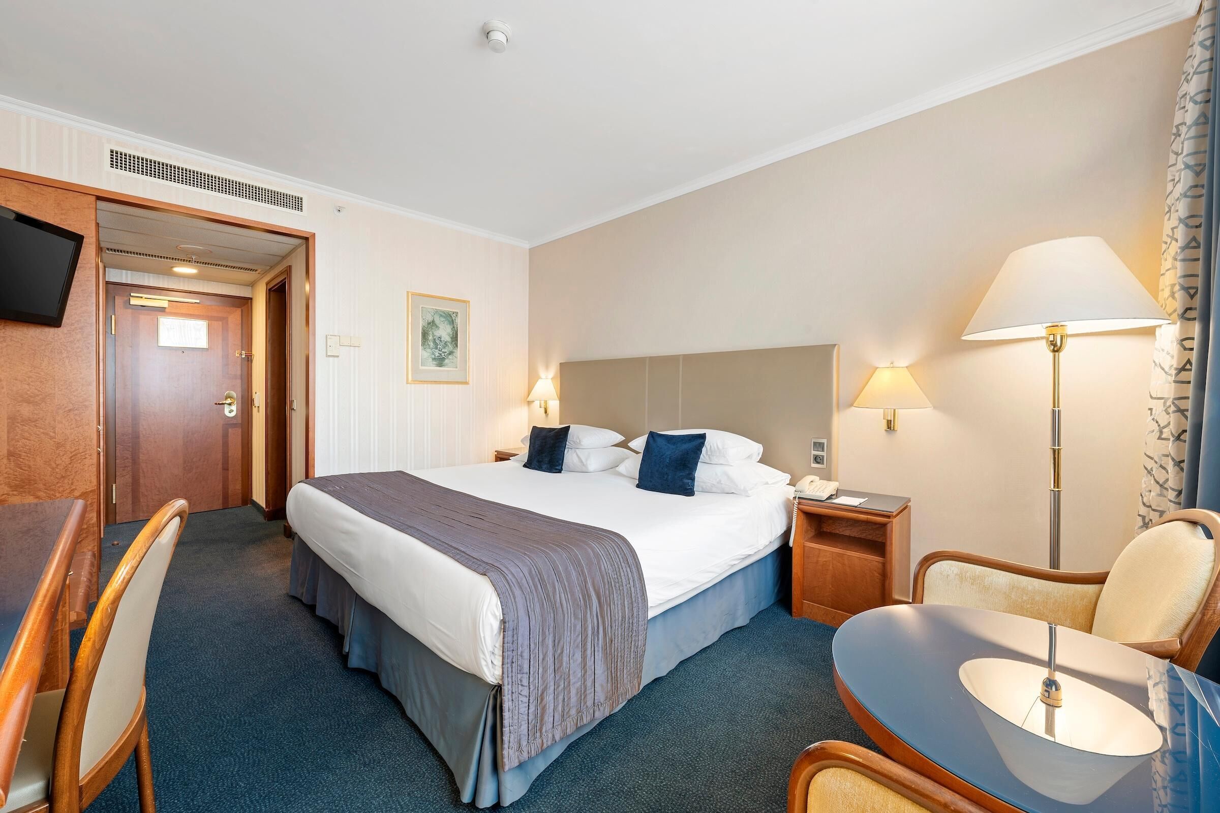 A hotel room with a bed, bedside tables, a desk, a chair, and a TV.