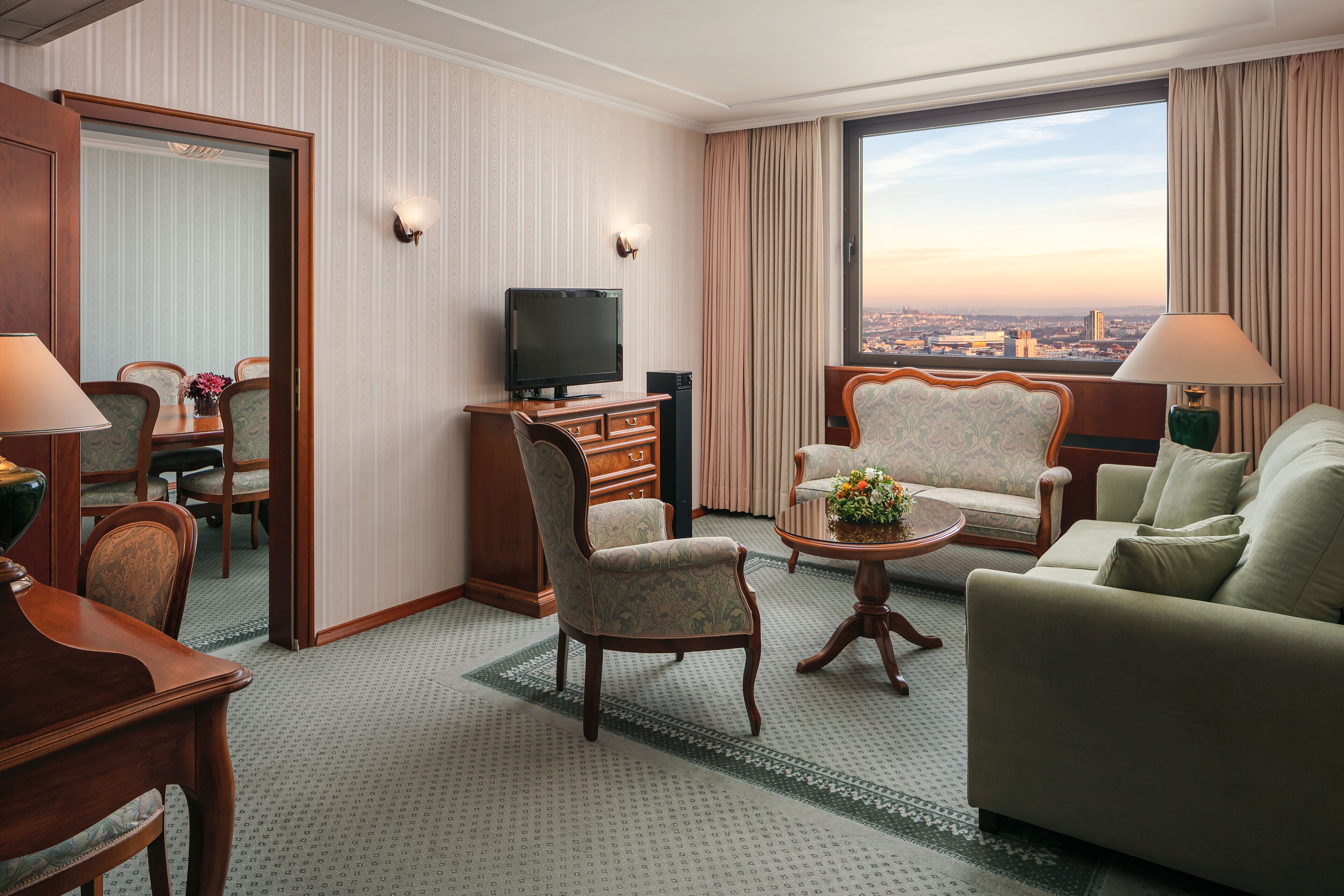 A hotel room with a dining area, a television, a sofa, and a view of the city.