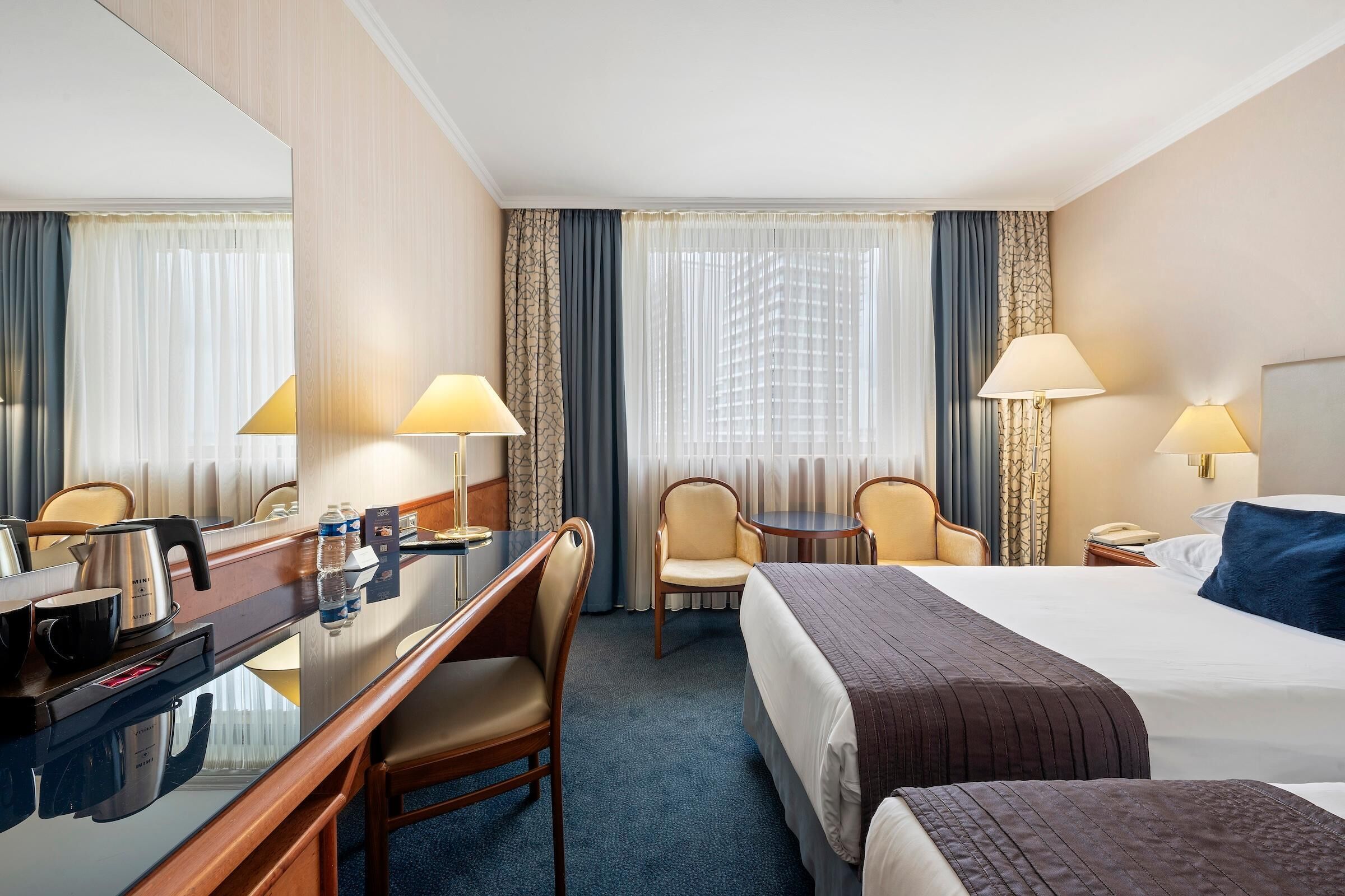 A hotel room with a bed, a desk with a chair, a small table, and two lamps.