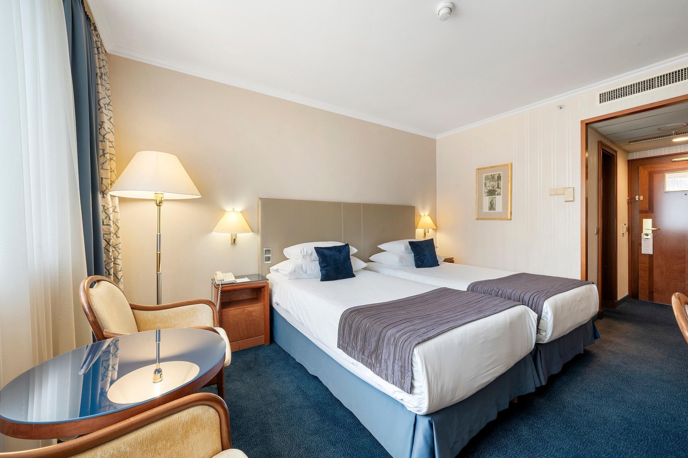 Ein Hotelzimmer mit zwei Betten, einem Schreibtisch, einem Stuhl, einer Lampe und einer Tür.