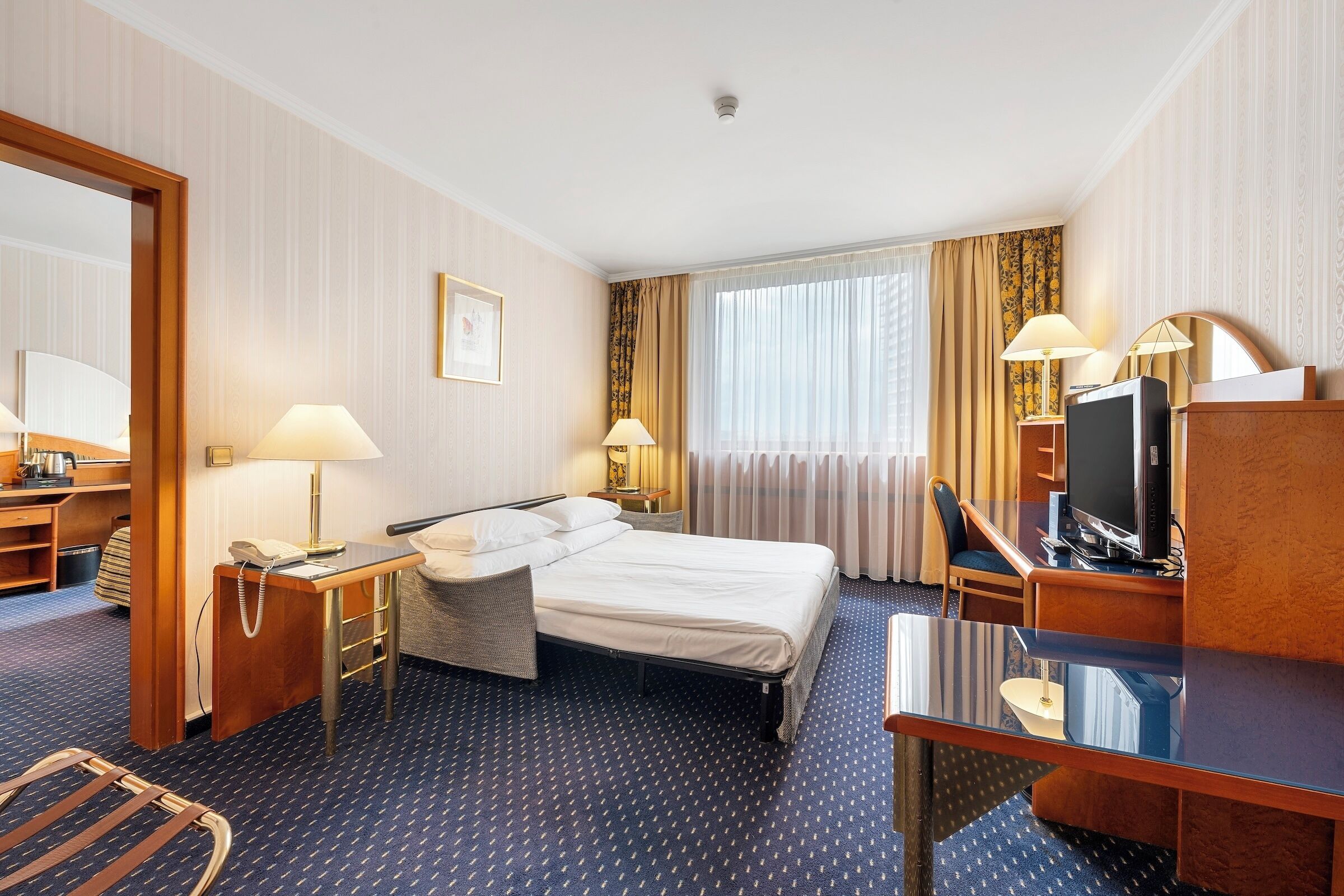 Habitación de hotel con cama, escritorio, televisor y ventana con cortinas.