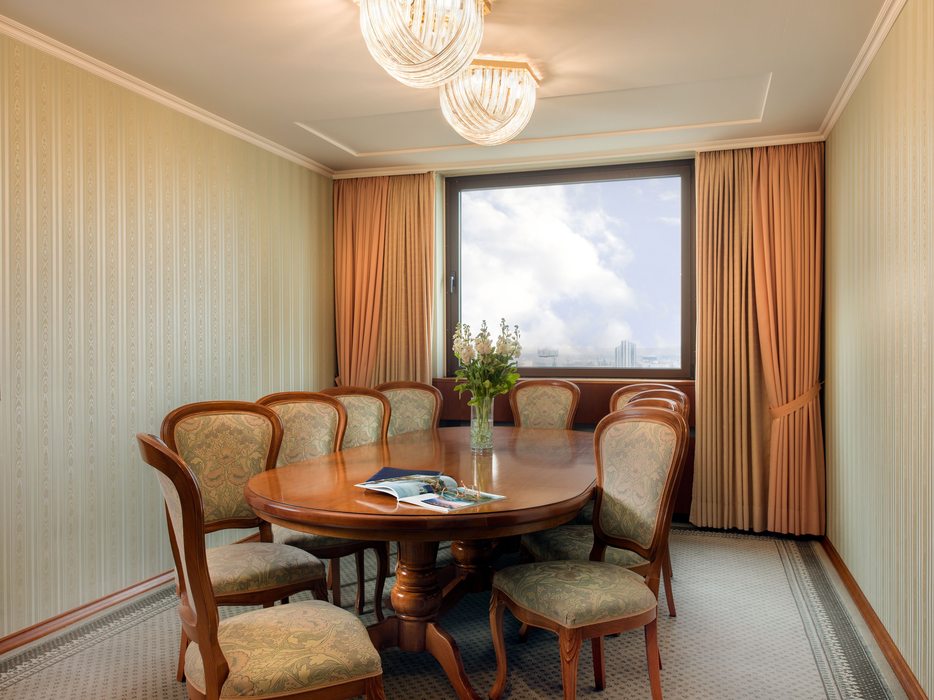 A conference room with a round table, eight chairs, a vase with flowers, and a large window with curtains.