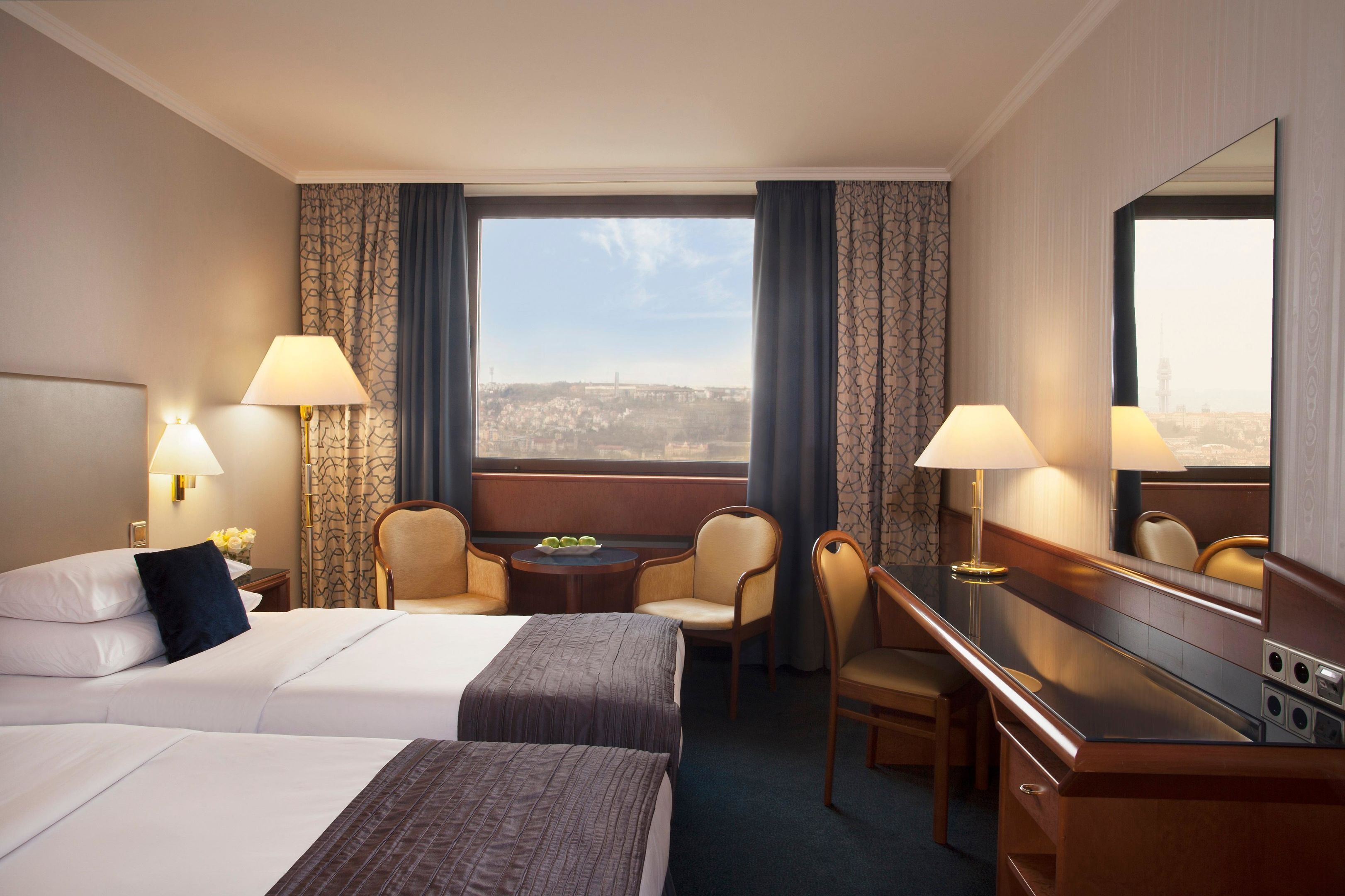 A hotel room with a large bed, two chairs, a desk, a mirror, and a view of a cityscape.