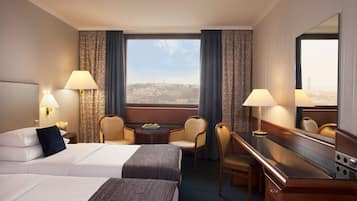 A hotel room with a large bed, two chairs, a desk, a mirror, and a view of a cityscape.