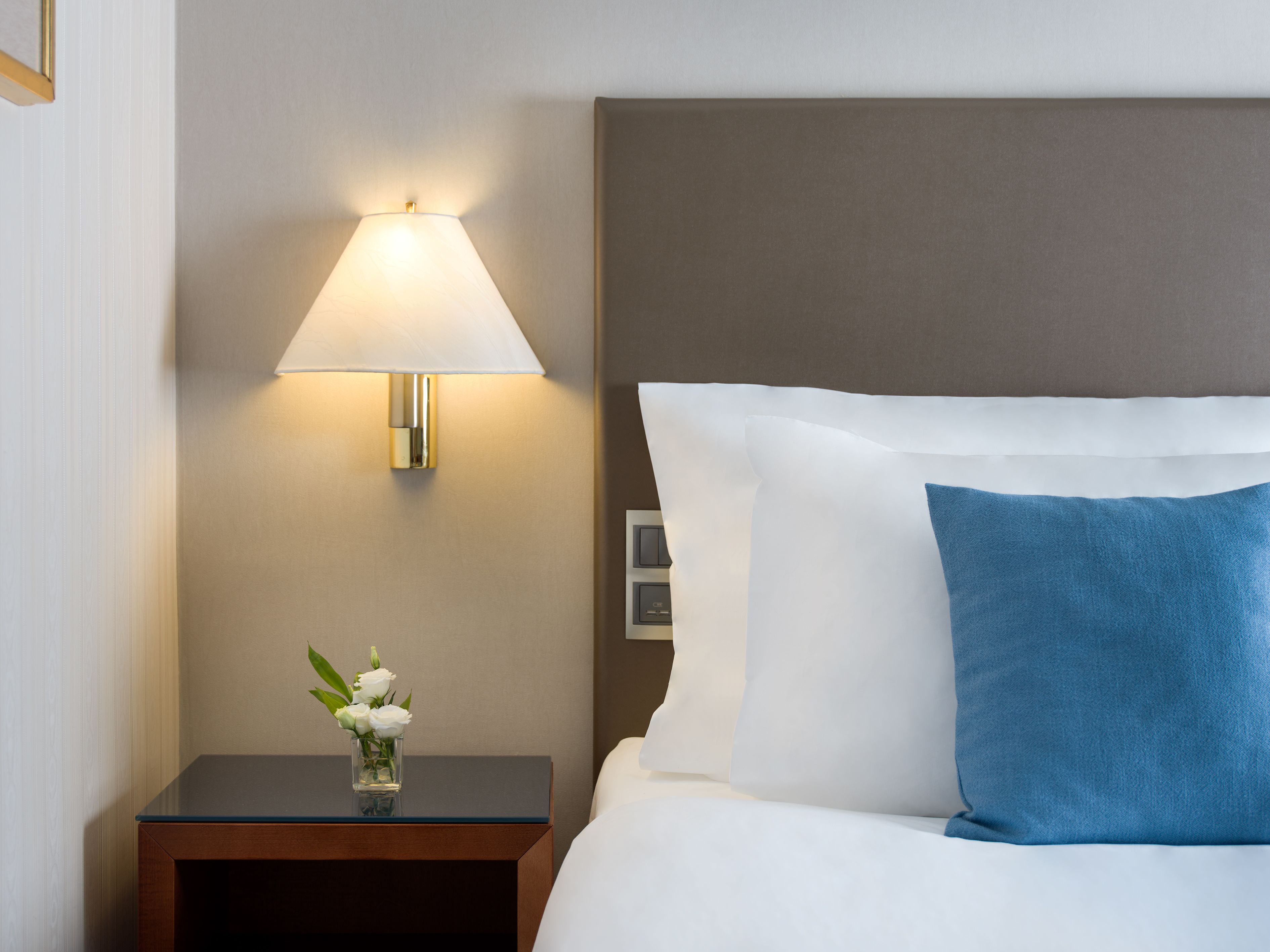 Una cama bien hecha con almohadas blancas y azules, una lámpara de noche y un pequeño jarrón con flores blancas sobre una mesita de madera.