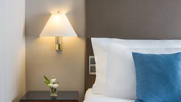 Una cama bien hecha con almohadas blancas y azules, una lámpara de noche y un pequeño jarrón con flores blancas sobre una mesita de madera.