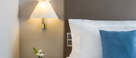 Una cama bien hecha con almohadas blancas y azules, una lámpara de noche y un pequeño jarrón con flores blancas sobre una mesita de madera.