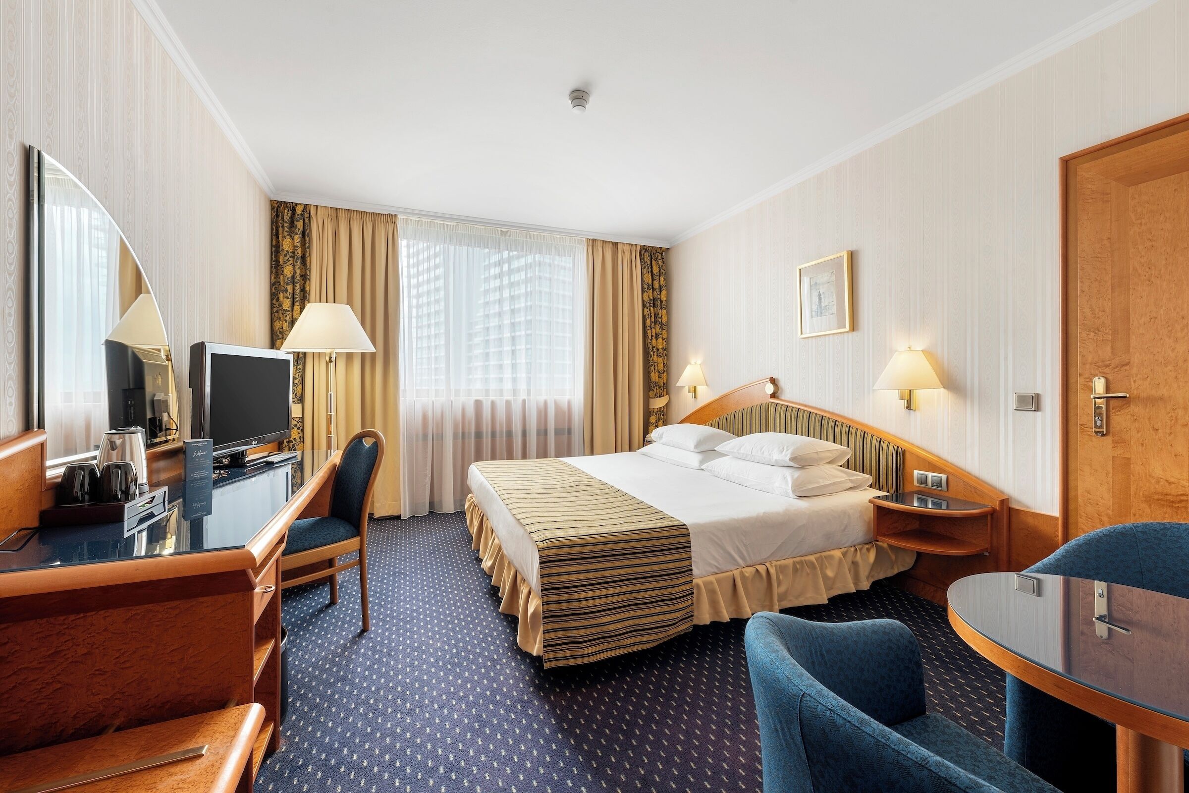 A hotel room with a bed, a desk with a television, a chair, a table, and a large mirror.