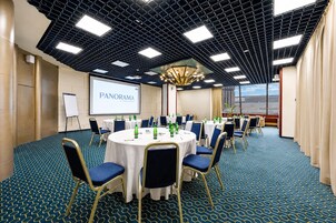 Een congrescentrum met ronde tafels, stoelen, een projectiescherm en een groot raam.
