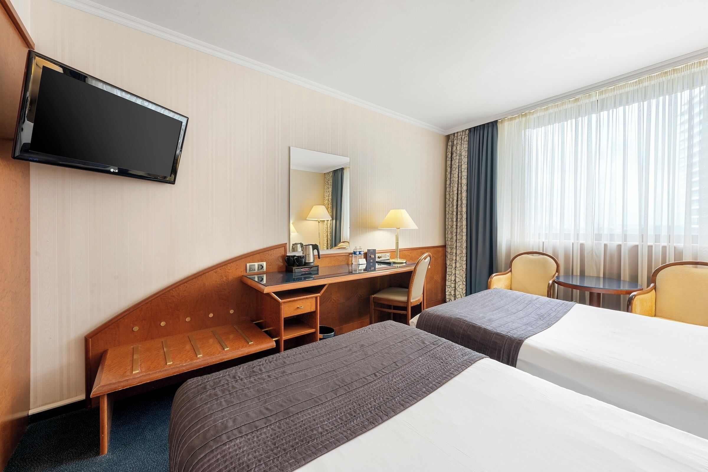 Een hotelkamer met twee bedden, een bureau met stoel, een televisietoestel en een groot raam met gordijnen.