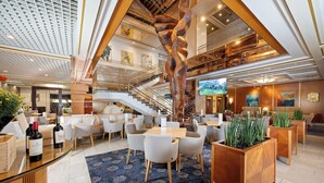A spacious hotel lobby with a central staircase, multiple seating areas, and a large screen displaying a scenic view. - Panorama by Verdi Hotels (Prague)