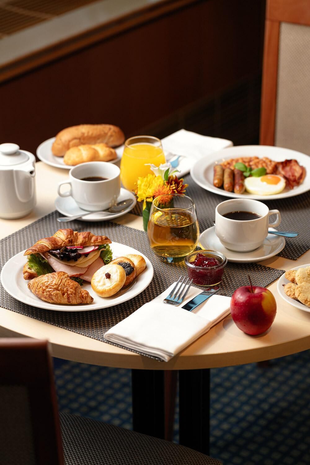A breakfast table with croissants, a sandwich, a croissant with a pat of butter, a glass of orange juice, a cup of coffee, a glass of tea, a plate with bacon, eggs, and a red apple.