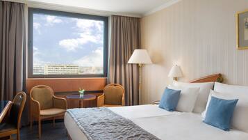 Una habitación de hotel con una cama grande, dos sillas, una mesita, una lámpara y vistas a la ciudad a través de un amplio ventanal.