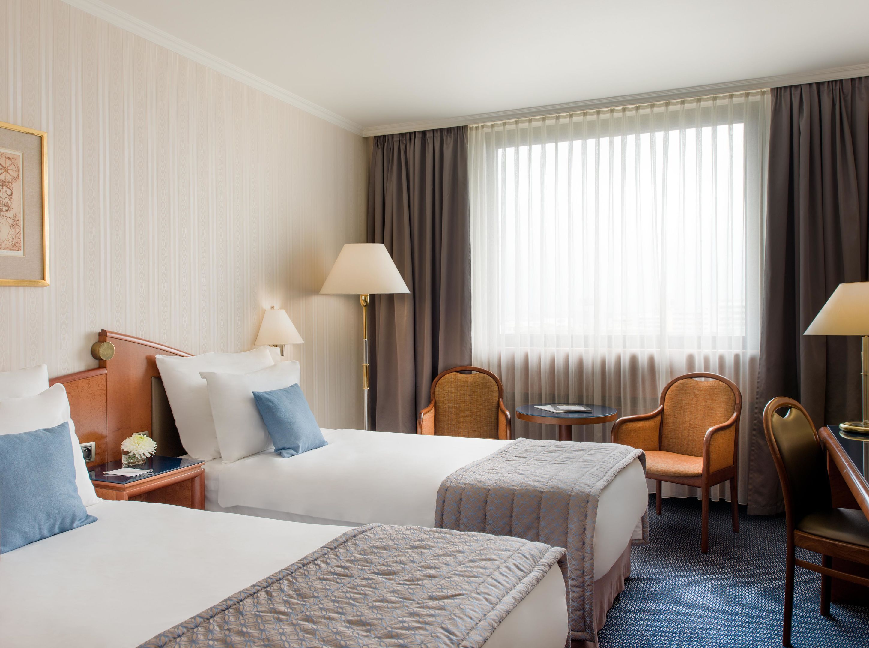 A hotel room with two beds, a small table, two chairs, a lamp, and a window with curtains.