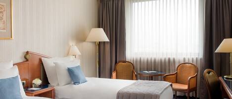A hotel room with two beds, a small table, two chairs, a lamp, and a window with curtains.