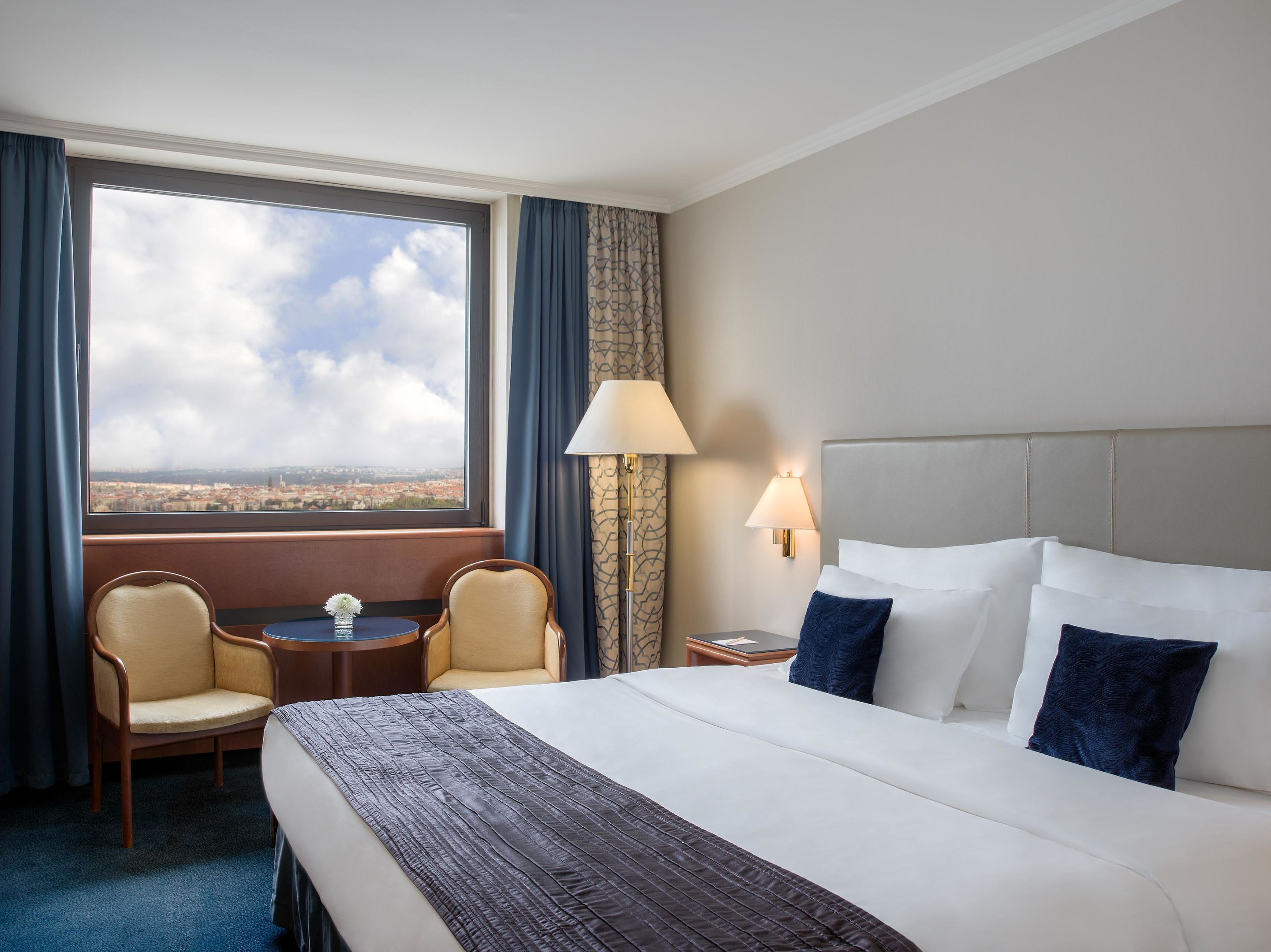 A hotel room with a large bed, two armchairs, a small table, a lamp, and a view of the city through a large window.