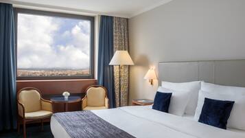 A hotel room with a large bed, two armchairs, a small table, a lamp, and a view of the city through a large window.