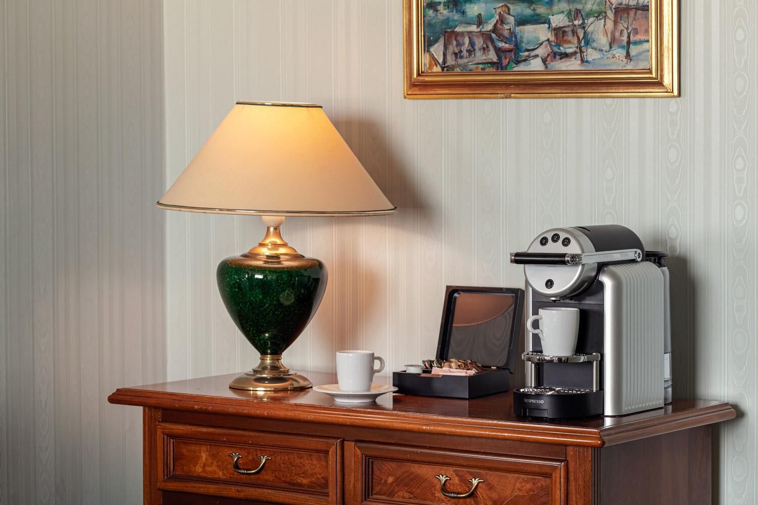 Et træbord med en grøn lampe, en hvid kop, et indrammet billede og en kaffemaskine.
