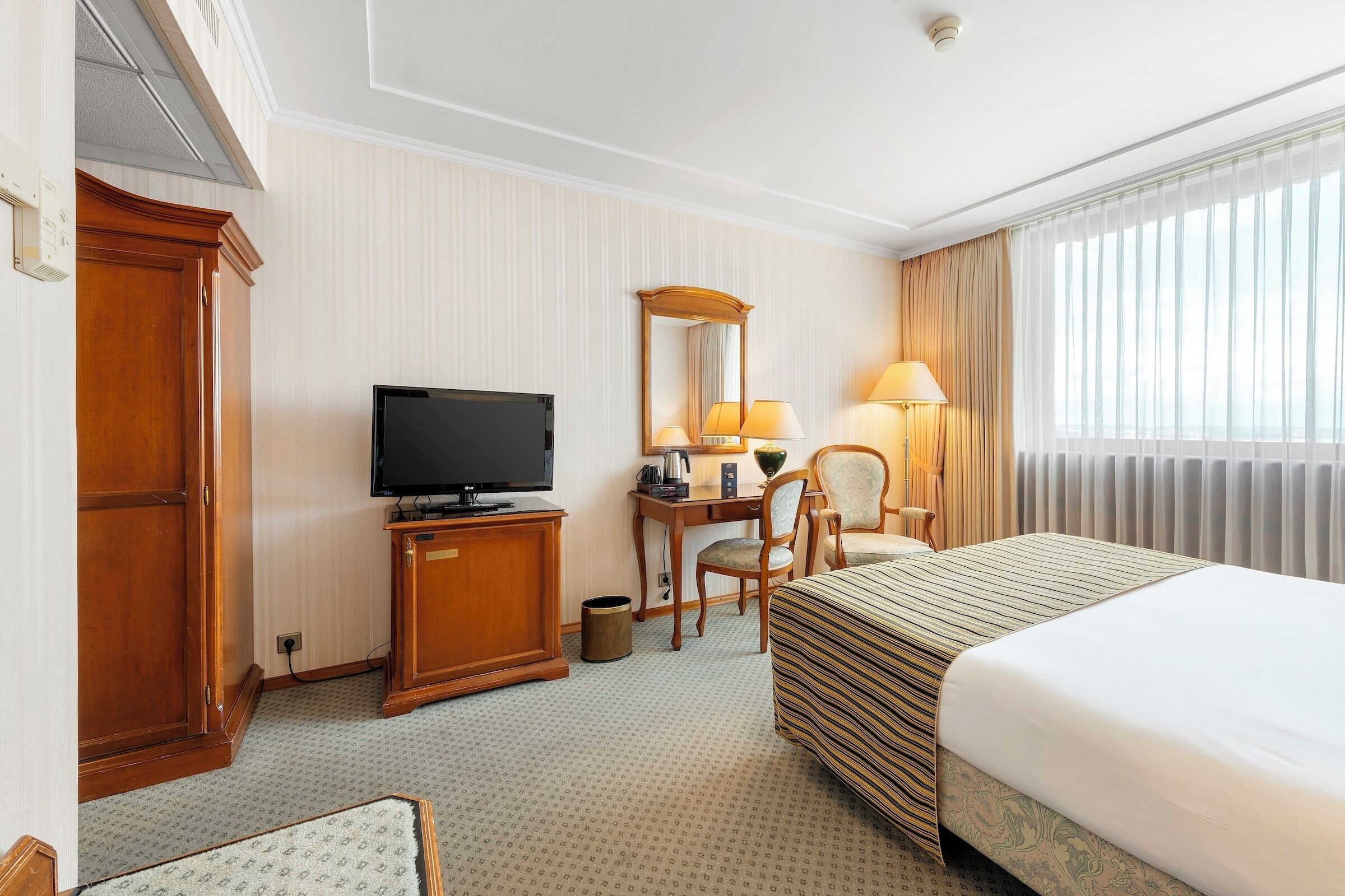 Una habitación de hotel con cama, escritorio con televisor, silla y ventana grande con cortinas.