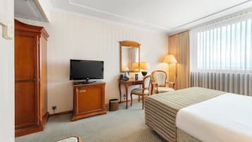 Una habitación de hotel con cama, escritorio con televisor, silla y ventana grande con cortinas.