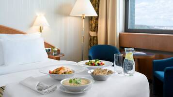 Hotelkamer met een bed, een eettafel met eten, een lamp, een stoel en uitzicht op de stad.
