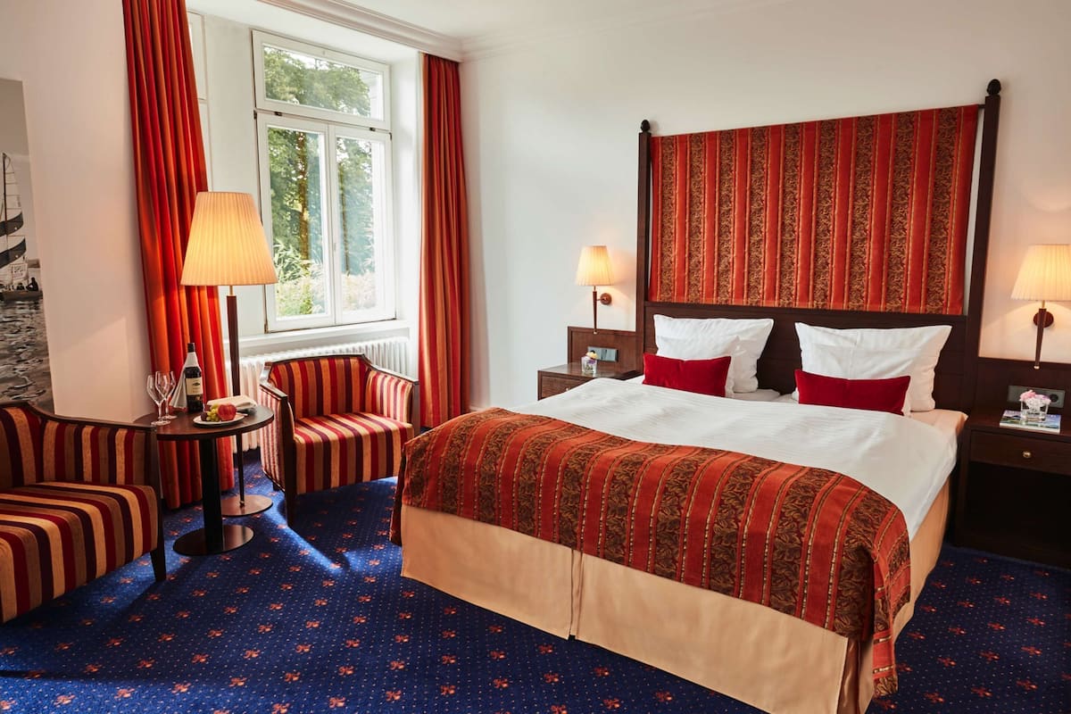 Une chambre d’hôtel avec un grand lit, deux tables de chevet, une chaise, une petite table et une fenêtre avec des rideaux.