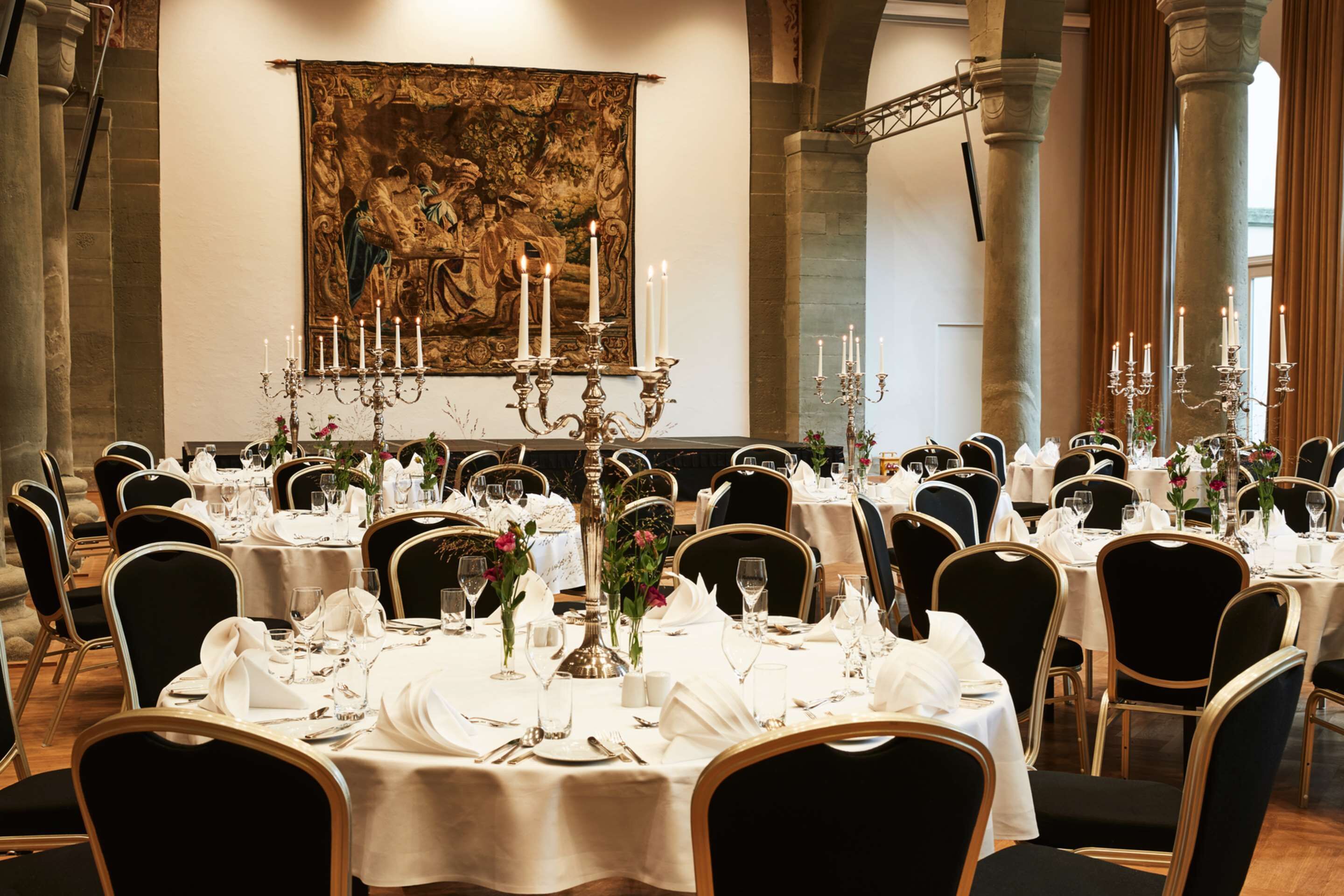 Ein Festsaal mit runden Tischen, gedeckt für ein Essen, mit weißen Tischdecken, schwarzen Stühlen und Kerzenlüstern.