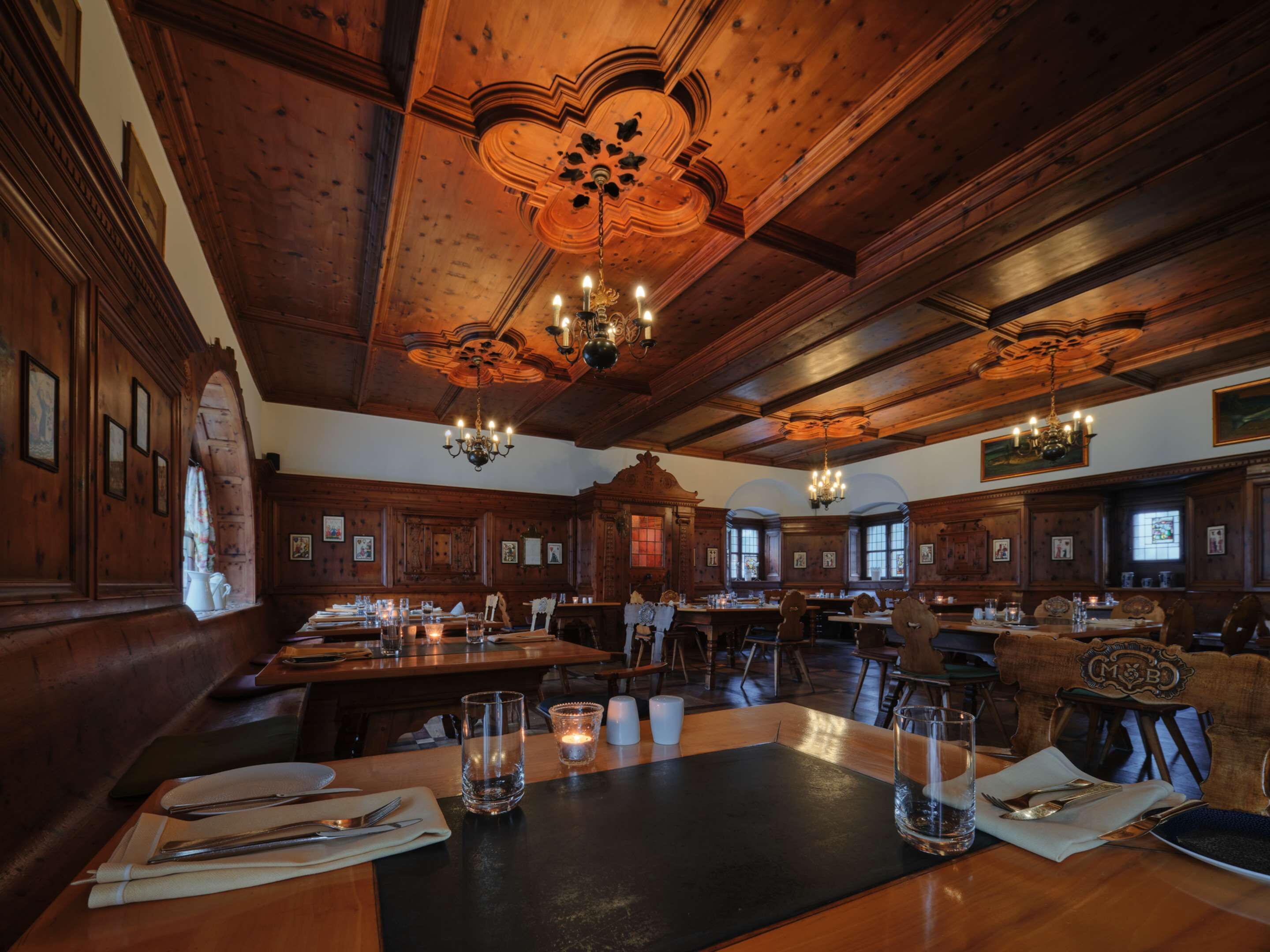 Une salle à manger spacieuse, avec des boiseries et des lustres. Les tables sont dressées avec des couverts et des verres.